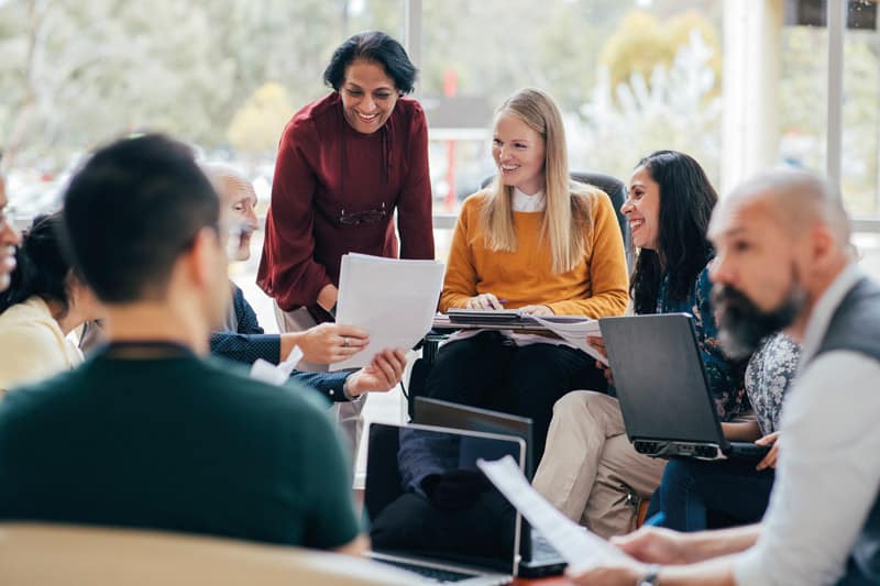 A diverse team looks happy in conversation. They are sitting around with laptops in front of them. Woman of Indian ethnicity leads the team